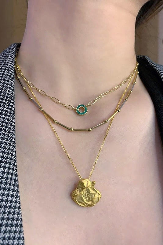 A close-up of a woman's chest and neck, showcasing three layered gold necklaces. The shortest is a delicate chain with a small green malachite circle pendant. The middle features a chain with small gold bar and bead accents. The longest has a textured, abstract gold pendant. The woman is wearing a gray and black plaid blazer.