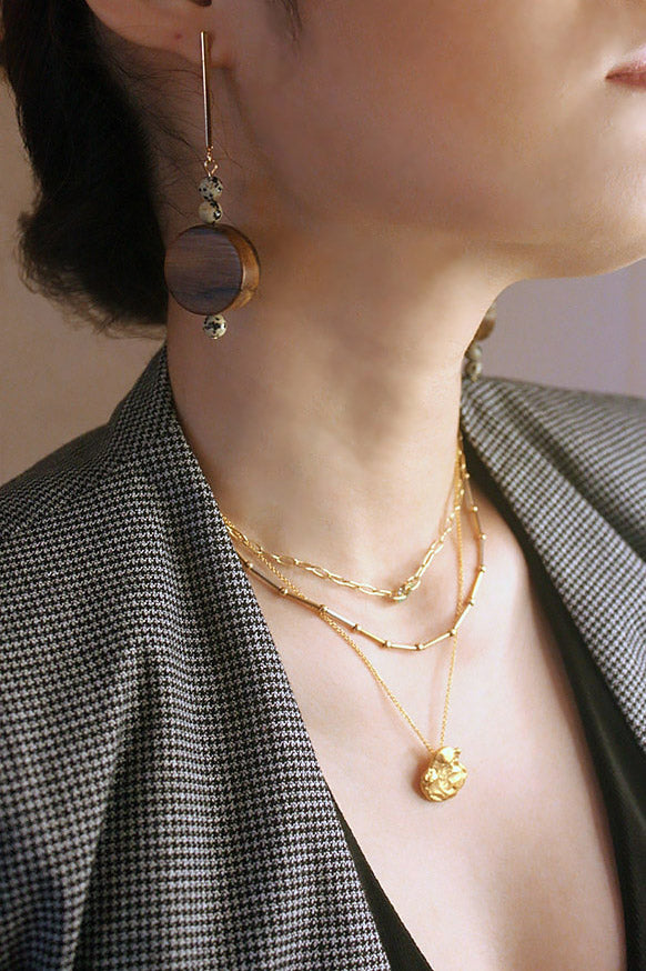 A close-up side profile of a woman's ear and chest, showcasing a long, dangling earring with a dark wood disc and spotted beads. The woman is also wearing three layered gold necklaces: a paperclip chain with a malachite ring, a beaded bar chain, and a delicate chain with an abstract gold pendant. The woman is wearing a grey and black plaid blazer.