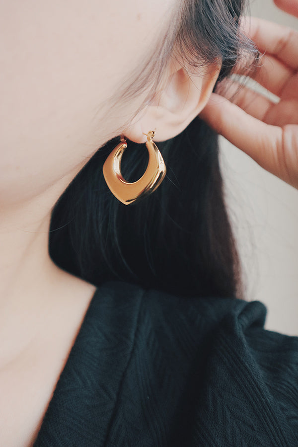 A close-up image of a womans ear wearing a bold, gold square hoop earring with a smooth, polished finish. The earring has a geometric design that gives it a modern, sophisticated look, perfect for making a statement.