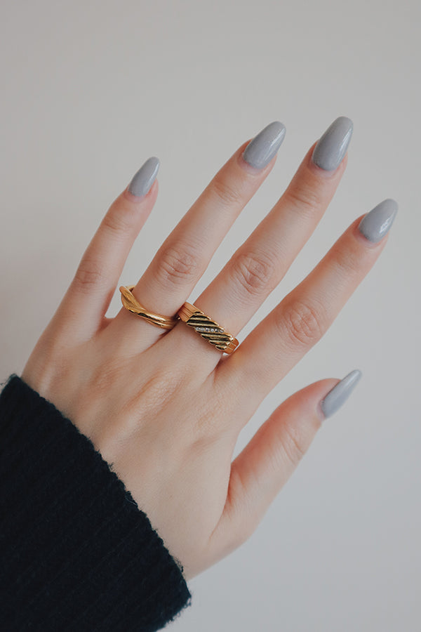 A female hand with long, elegant grey manicured nails, wearing two gold rings on the ring finger: one is a sleek, wavy band, and the other is a wider, textured band with a diagonal line of small, sparkling clear stones. The woman is wearing a black long-sleeved top.
