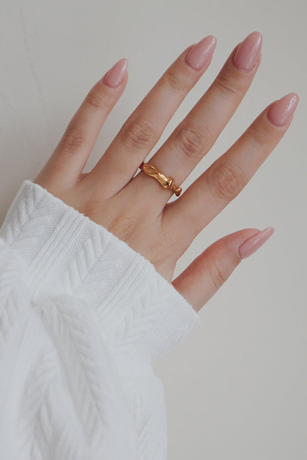 A detailed image of a womans hand wearing an irregularly shaped gold-plated ring. The ring features a wavy, organic design that adds a touch of modern elegance, making it a stylish accessory for any occasion.