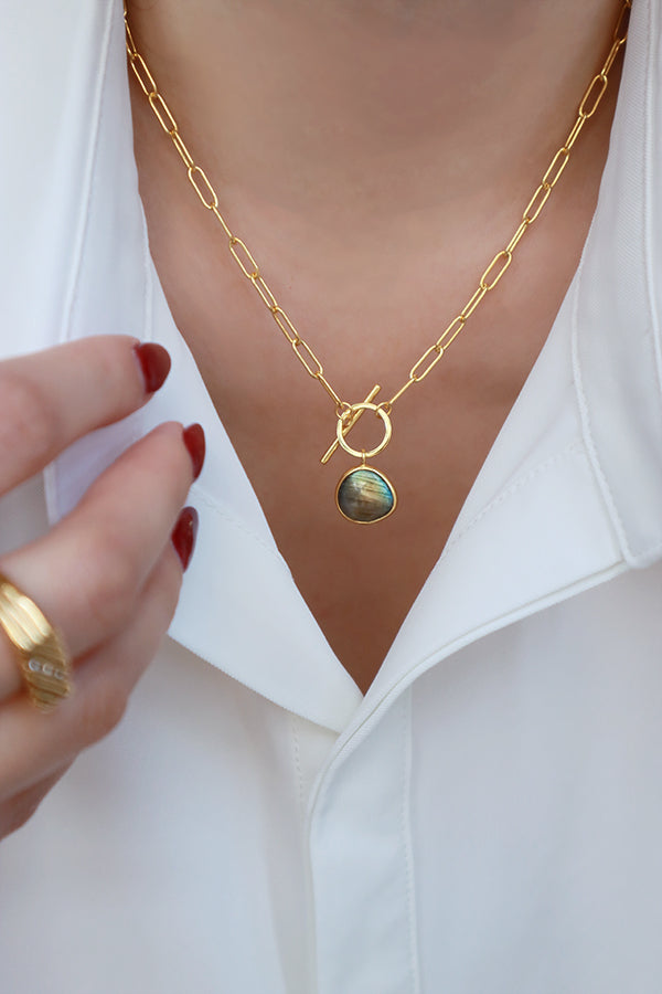 A woman's neck wearing a trendy gold paperclip chain necklace with a toggle clasp and a beautiful iridescent labradorite gemstone pendant, paired with a white shirt.