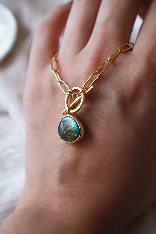 A woman's hand holding a stylish gold paperclip chain necklace with a unique labradorite gemstone pendant and toggle clasp, highlighting its design.