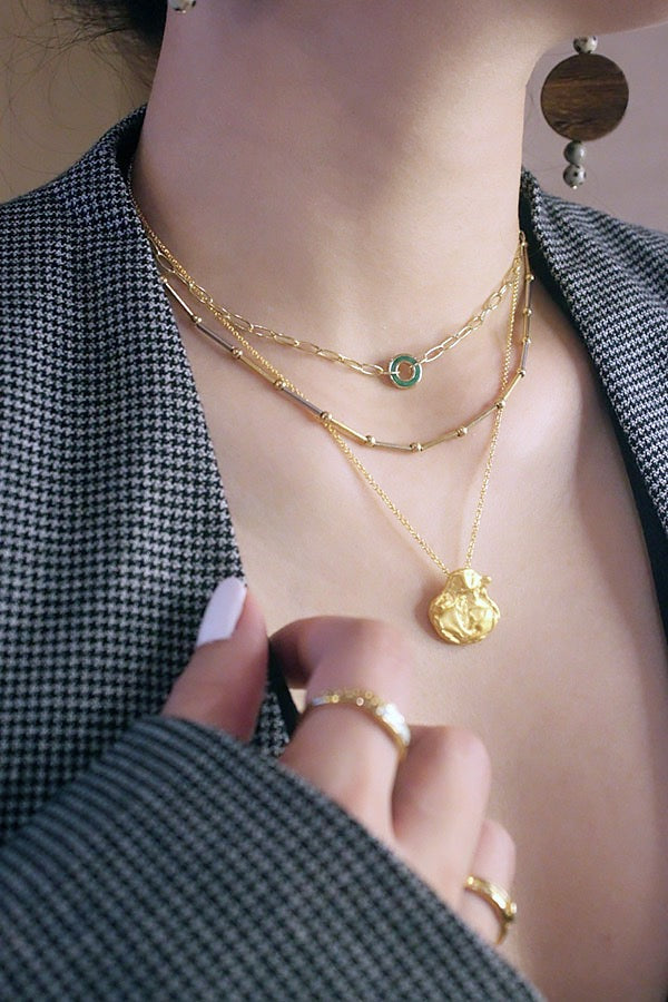 A close-up of a woman's chest and neck, showcasing three layered gold necklaces. The shortest is a delicate chain with a small green malachite circle pendant. The middle features a chain with small gold bar and bead accents. The longest has a textured, abstract gold pendant. The woman is wearing a gray and black plaid blazer, and large, round, wooden earrings with spotted details are visible on her ears. Her hand, with white manicured nails and gold rings, is touching her collar.