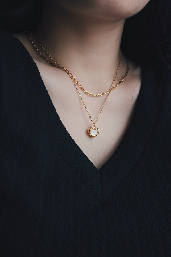 Layered gold necklace set featuring a heart-shaped pendant with a mother-of-pearl inlay, worn with a black V-neck top. The necklace is styled with a second chain that adds dimension to the look.