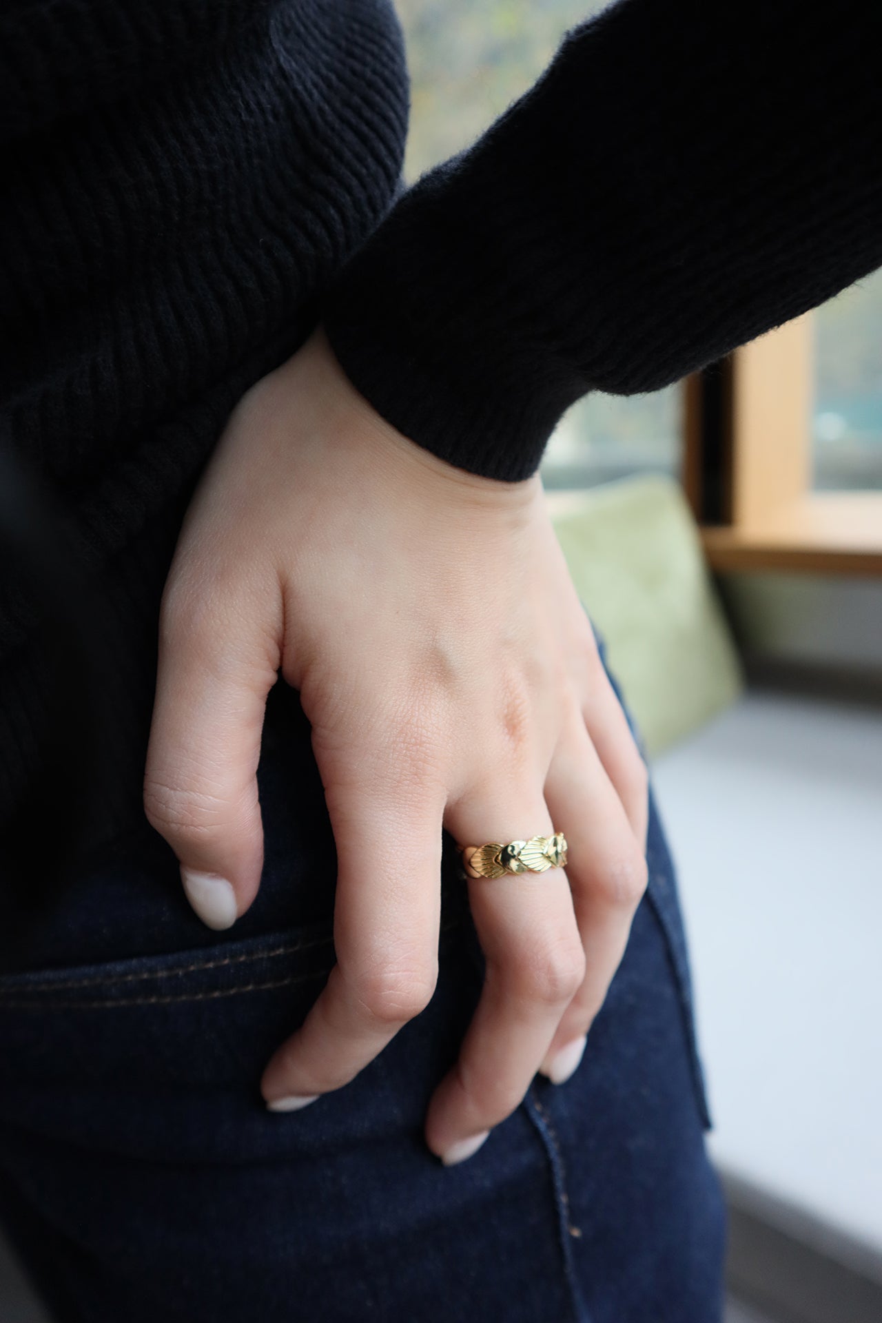 A female hand resting near a dark denim-clad leg, wearing a wide gold ring with a textured fan or shell design on her ring finger. The woman is wearing a black long-sleeved top and dark jeans.