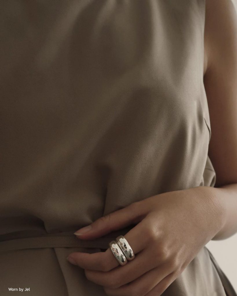 A close-up of a woman's hand, wearing two shiny silver stacked rings on her ring finger. The woman is wearing a sleeveless beige or light brown top, with a textured fabric. The bottom left corner states "Worn by Jet."
