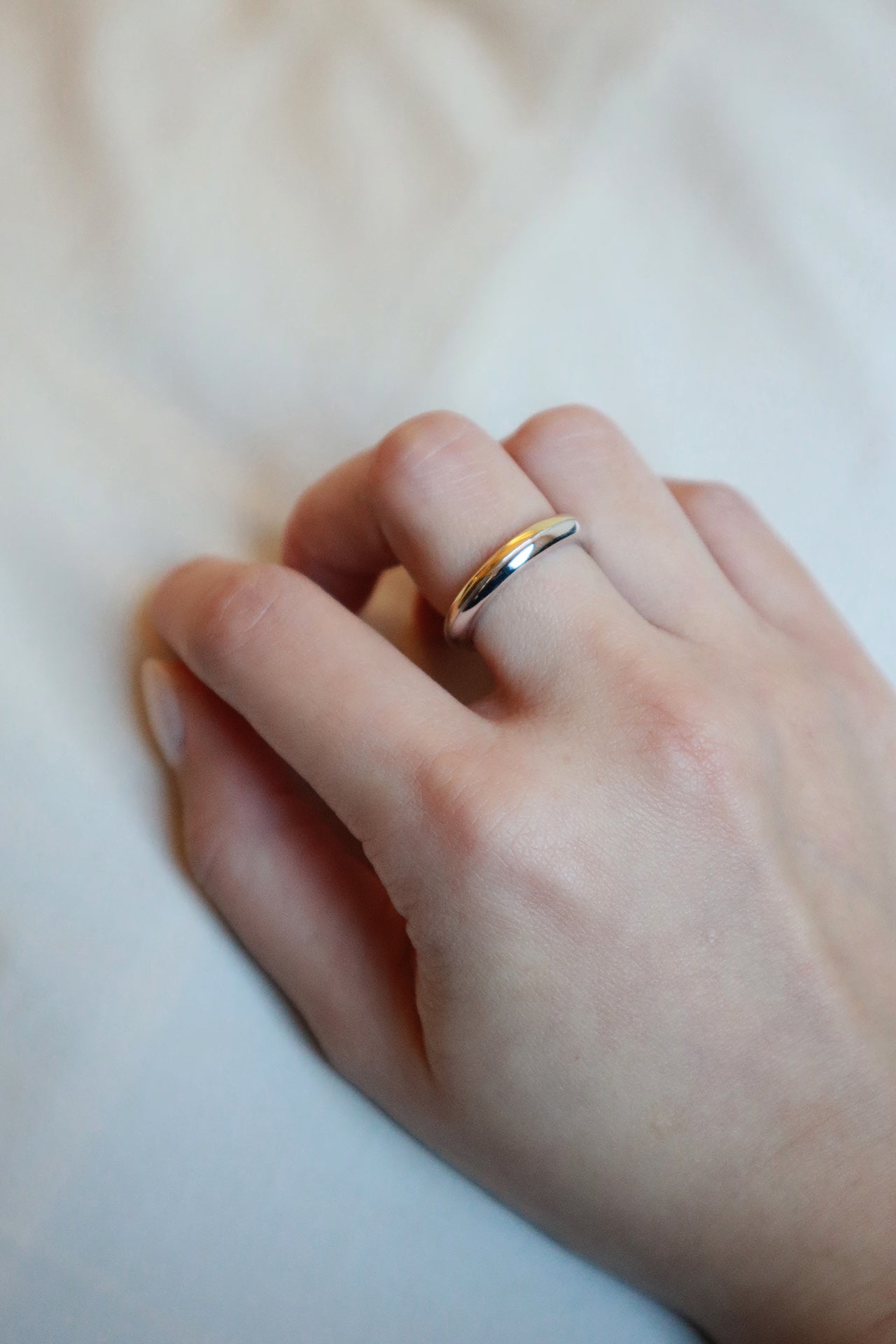 A close-up of a female hand, wearing a sleek, two-tone ring with a silver exterior and a gold interior on her ring finger. The background is a crumpled, light-colored fabric.