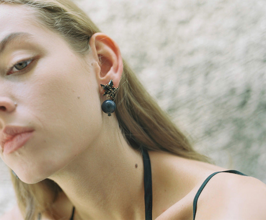 A close-up of a young woman with long, light brown hair, looking slightly to the side, showcasing an ornate black star-shaped stud earring from which hangs a spherical dark blue stone. The woman has a nose piercing and is wearing a dark strap top, against a blurred light-colored textured background.