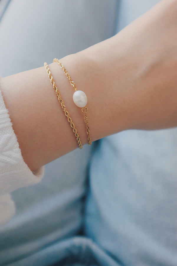 手腕上佩戴兩條疊戴金色手鍊:一條有紋理的麻花鍊,和一條鑲有一顆白色珍珠的細緻鍊。