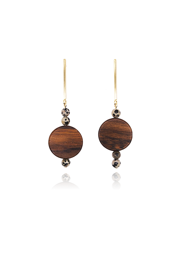 A standalone image of a pair of long, dangling gold earrings. Each earring features a thin gold bar, a spotted dalmatian jasper bead, a large circular dark wood disc, and another spotted dalmatian jasper bead at the bottom. The earrings are displayed vertically on a black background with reflections below.