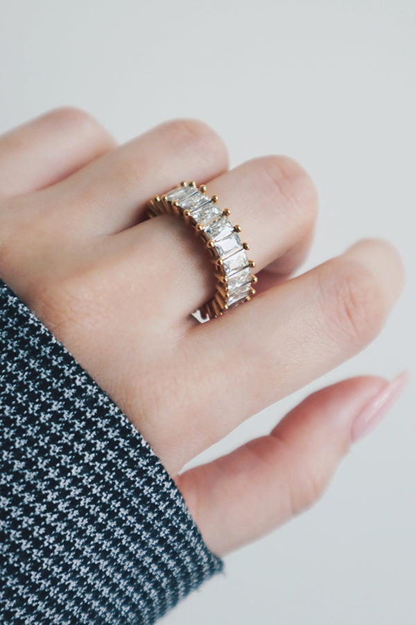 A side view of a hand wearing a gold eternity ring with a row of rectangular-cut cubic zirconia stones. The rings substantial design and brilliant stones create a bold and glamorous statement. The soft, textured sleeve adds an element of comfort, balancing the ring's opulent style with a touch of casual elegance.