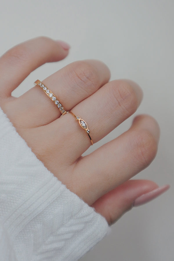 A hand wearing two delicate gold rings: one with a full band of small cubic zirconia stones, and another with a marquise-shaped center stone flanked by two round stones.