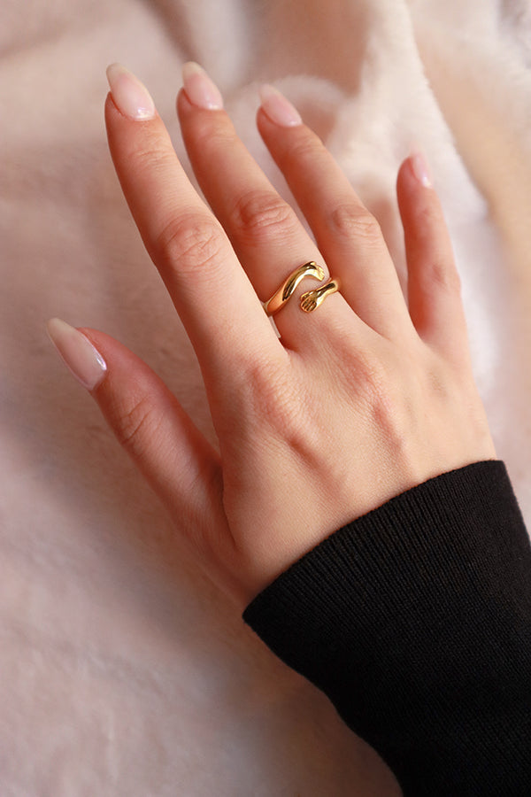 一隻女性手指佩戴著一枚閃亮的金色「擁抱」造型戒指,其設計巧妙地呈現兩臂或兩手溫柔環繞手指的姿態,造型別緻。