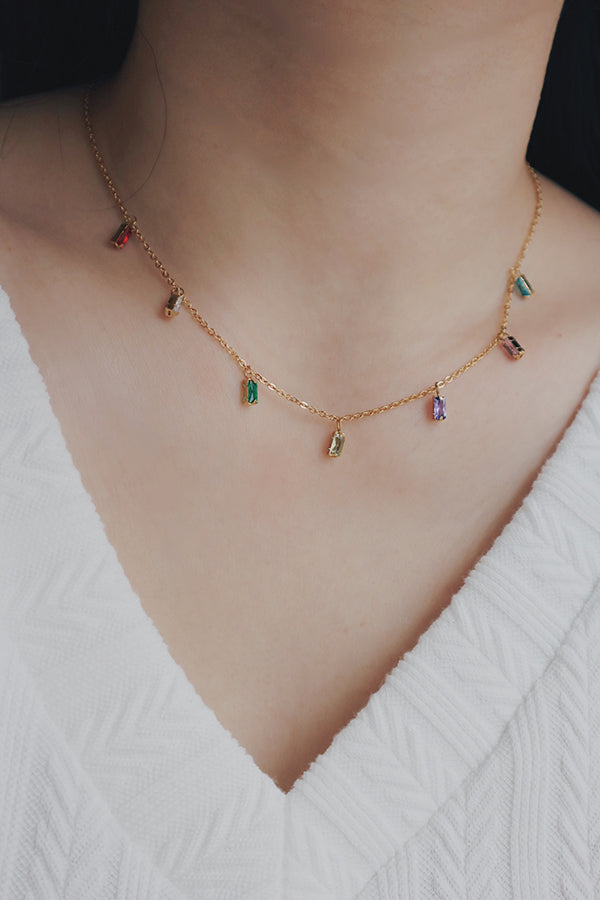 A detailed image showing a womans neckline adorned with a gold chain necklace, highlighted by small, colorful cubic zirconia stones hanging at intervals. The necklace exudes a cheerful and youthful vibe, making it a versatile accessory for various occasions.