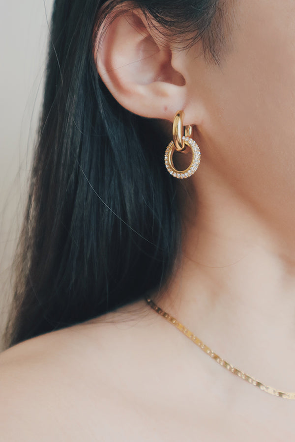 A detailed image of a womans ear adorned with gold hoop earrings that include a dangling charm covered in sparkling cubic zirconia stones. The earrings are designed to catch the light, adding a touch of glamour and sophistication to any outfit.