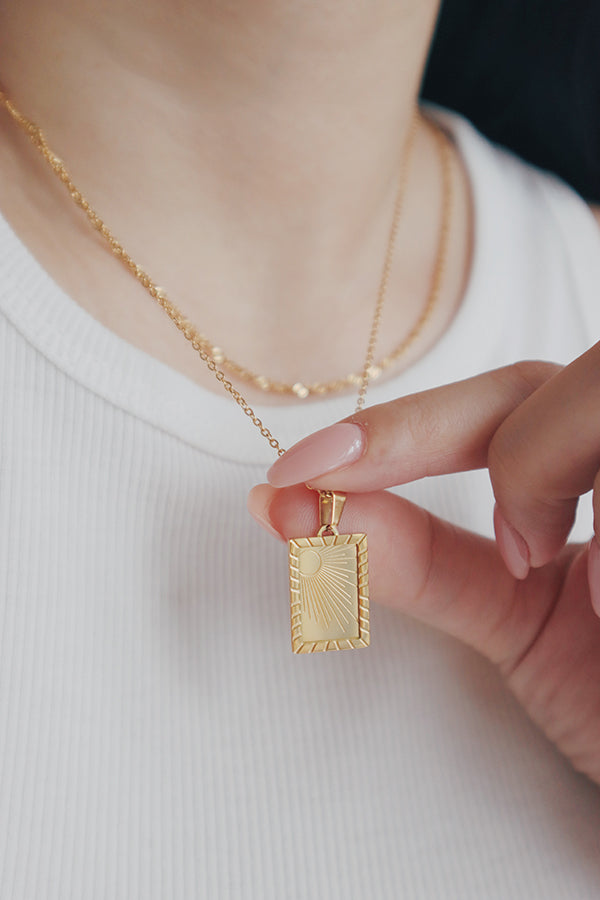 A woman wearing the SH & Co. Savita Sun necklace, featuring a rectangular pendant with an engraved sunburst design, paired with a gold chain, styled with a white textured V-neck top.