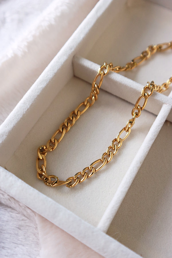 An overhead shot of a shiny gold Figaro chain necklace, neatly laid out inside a white velvet jewelry box. The necklace features the characteristic alternating pattern of one long and two short links.