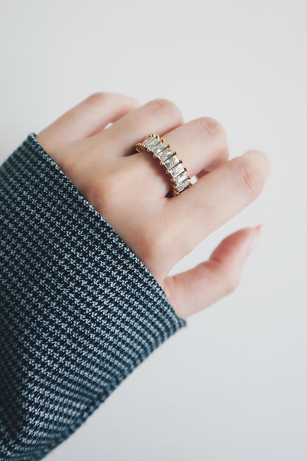 A close-up of a hand wearing a gold eternity ring encrusted with rectangular-cut cubic zirconia stones. The ring's wide band and dazzling stones catch the light beautifully, creating a stunning display of sparkle. The houndstooth-patterned sleeve adds a touch of modern style, complementing the ring's bold and glamorous design.