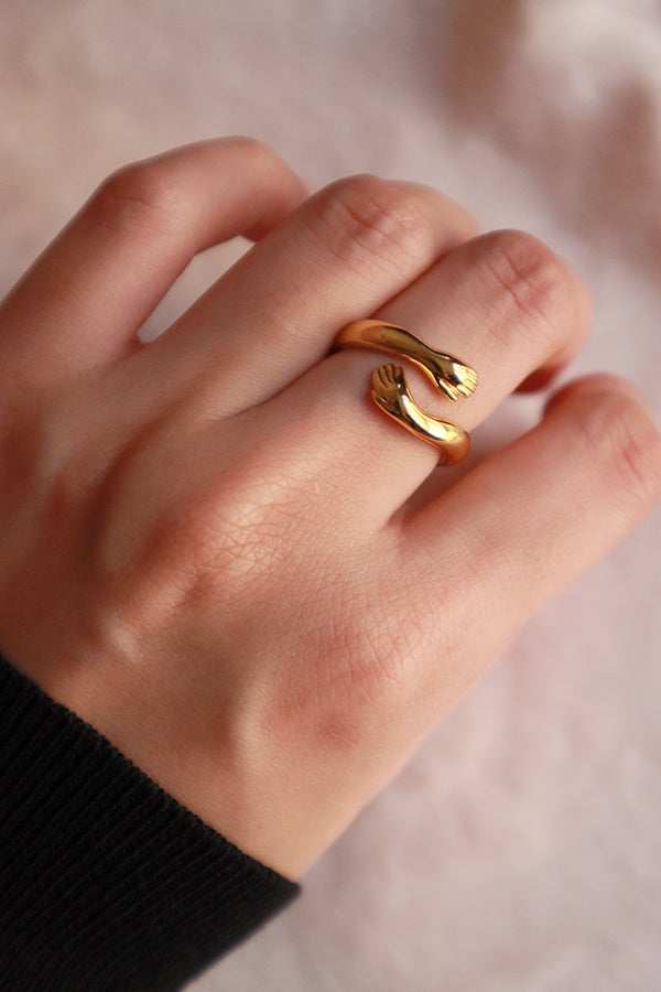 A woman's hand wearing a special gold ring designed as two stylized hands or arms meeting, forming a unique embrace shape on her finger.