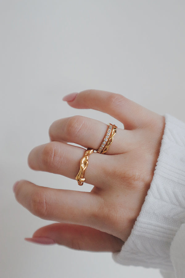 A close-up image of a womans hand wearing a set of gold rings, including an irregularly shaped ring with a fluid, organic design. The ring's uneven surface adds a unique texture, making it a standout piece in a stack of rings.