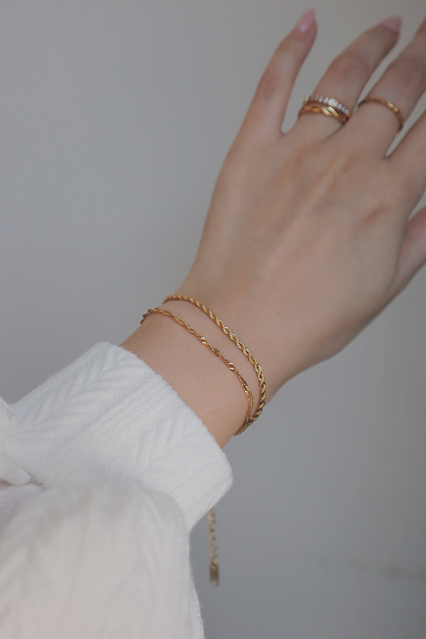 手腕上佩戴兩條疊戴金色麻花手鍊,背景中可見戴著金色戒指的手指。