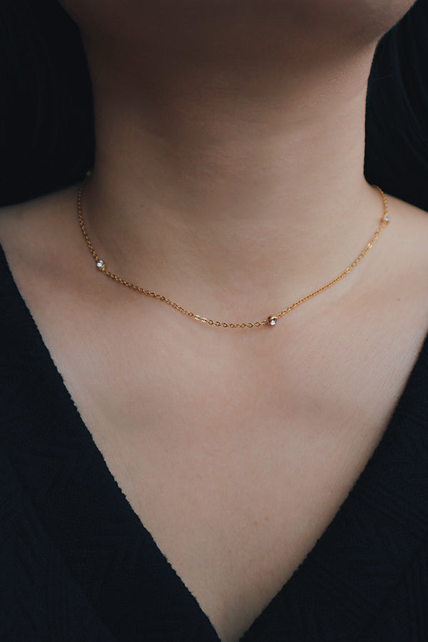 Minimalist gold necklace featuring small cubic zirconia stones evenly spaced along the chain, worn close to the neck for a choker-like appearance, paired with a black V-neck top.