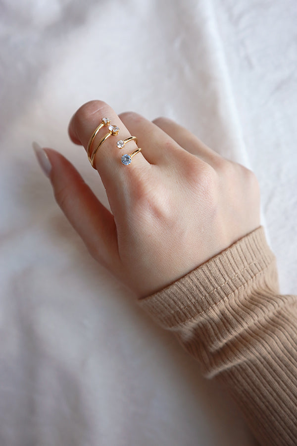 A hand wearing a unique gold fashion ring with a wrap-around design, featuring three sparkling cubic zirconia stones, one blue and two clear.
