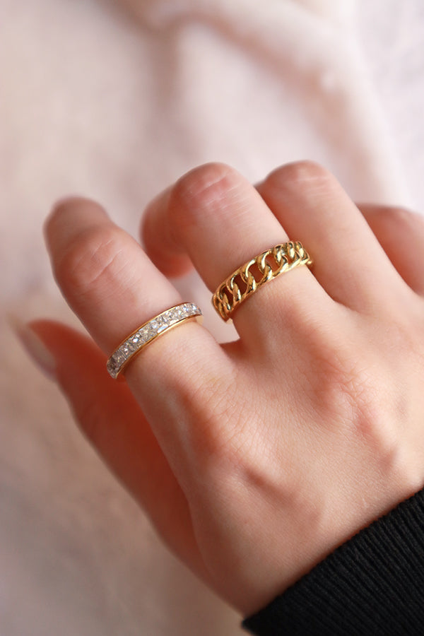 A female hand with fair skin, wearing two gold rings: a sparkling band of clear, rectangular-cut cubic zirconia stones on the middle finger, and a chunky Cuban link chain ring on the ring finger. The woman is wearing a dark long-sleeved top.