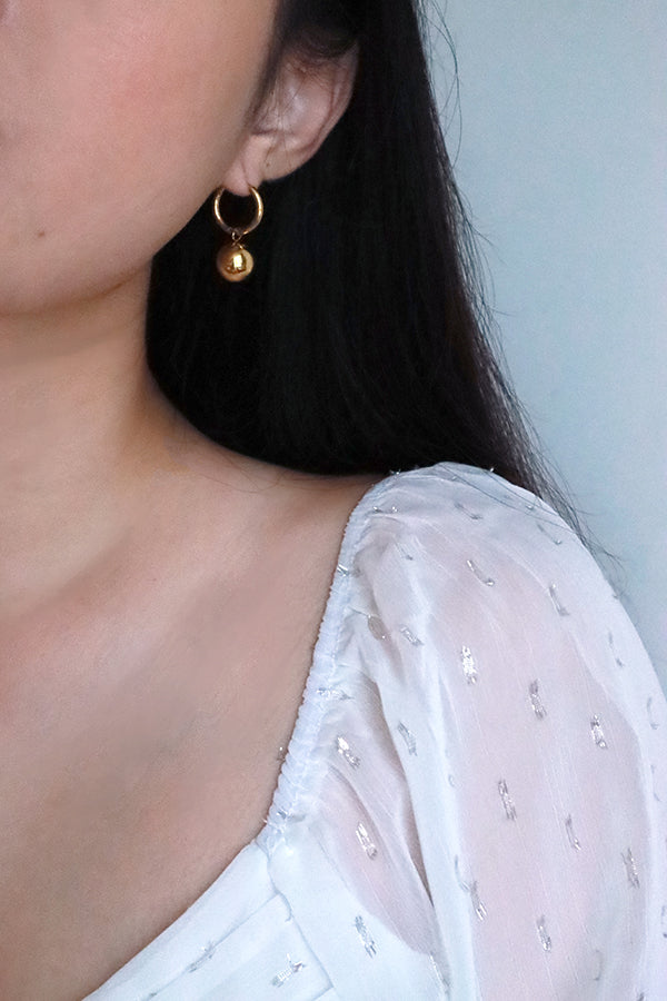 一隻耳朵佩戴著一款閃亮的金色圓圈耳環,下方懸掛著一顆拋光圓潤的金色球體,展現簡約日常風格。