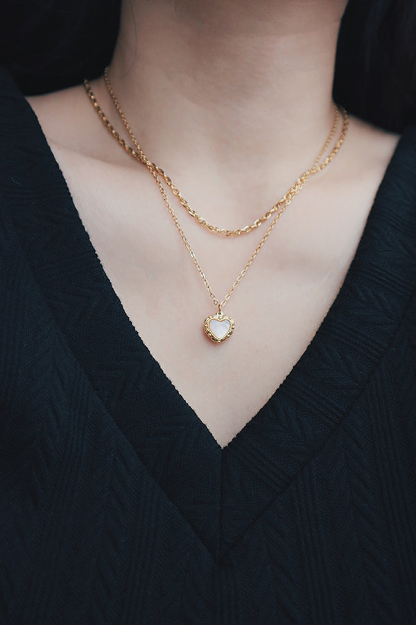 Close-up of a layered gold necklace set featuring a heart-shaped pendant with a mother-of-pearl inlay, worn with a black textured V-neck top.