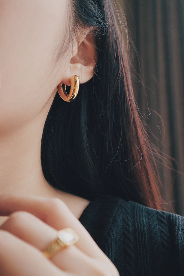 A close-up image of a womans ear wearing a pair of gold heart-shaped hoop earrings with a smooth, polished finish. The earrings have a bold, curved design that forms a heart shape, adding a playful and unique touch to her look.
