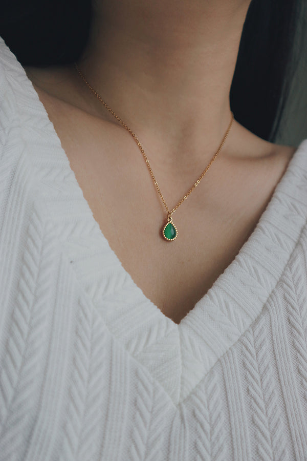 Close-up of a woman wearing a stainless steel waterproof necklace featuring a teardrop-shaped green gemstone pendant, styled with a white textured top.
