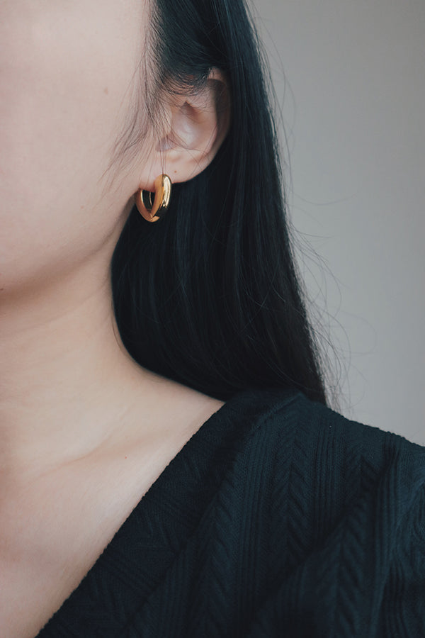 A detailed shot of a woman modeling gold heart-shaped hoop earrings. The earrings feature a sleek, heart-like shape with a polished, glossy surface, making them a fashionable and eye-catching accessory.