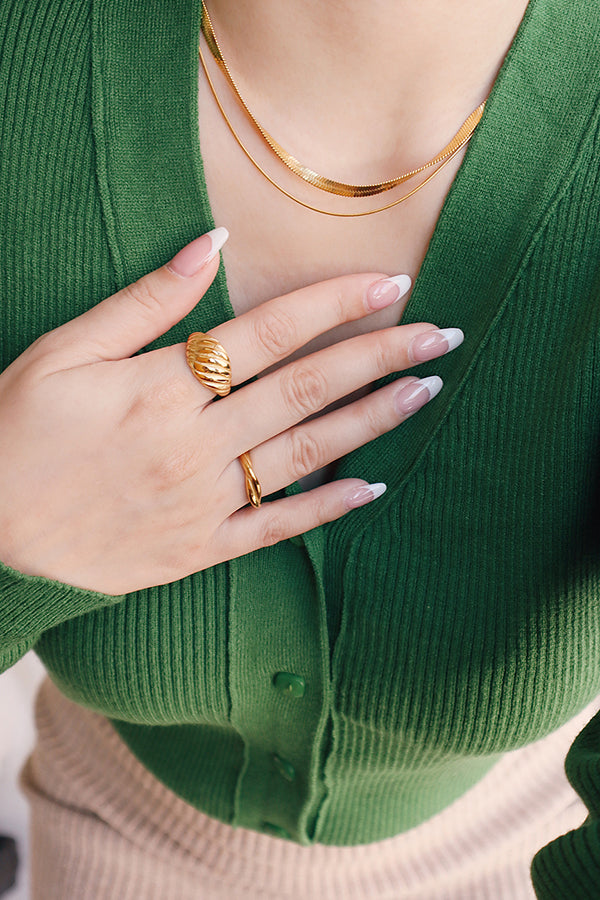 A woman's hand wearing a stylish chunky gold ring with a distinctive ribbed texture, paired with delicate layered gold necklaces, over a green ribbed top.