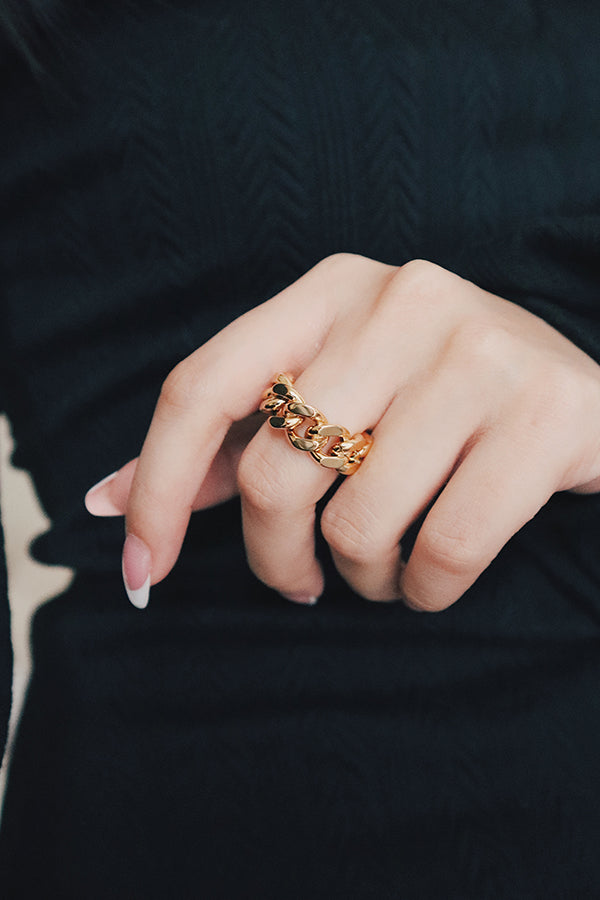 A person's hand with manicured nails, wearing a gold Cuban link chain ring on the ring finger. The person is wearing a dark, possibly black, garment. The ring appears robust and stylish.