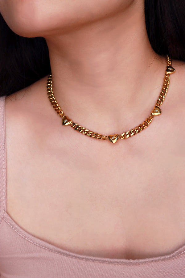 一位女性的頸部佩戴著一條不褪色的金色古巴鍊項鍊,鍊條上間隔鑲嵌著多個光滑拋光的心形墜飾,整體設計時尚且可愛。