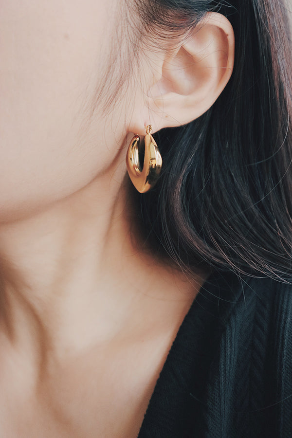 A detailed shot of a woman wearing a gold square hoop earring. The earring's unique square shape and shiny, smooth surface add a contemporary twist to the classic hoop style, making it an eye-catching accessory for any outfit.