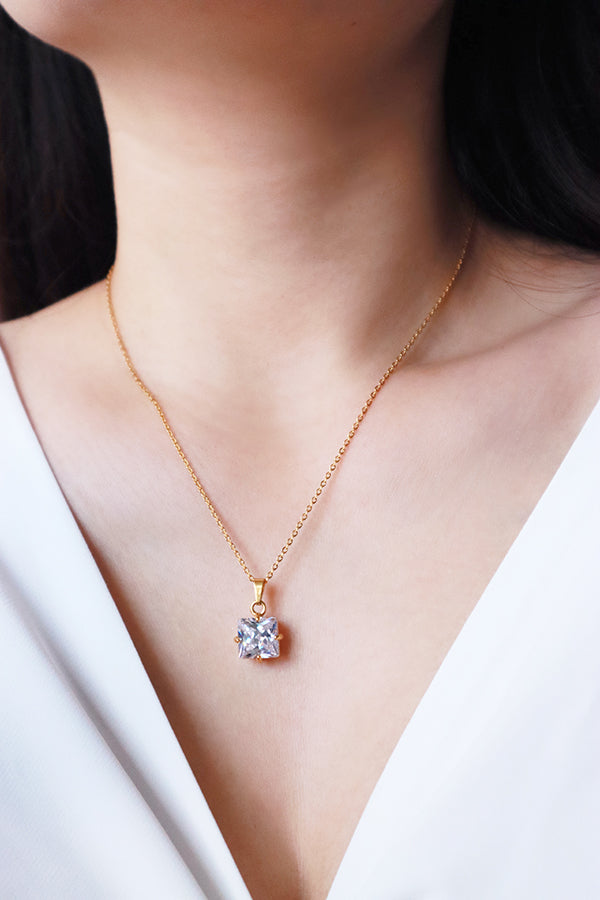 A close-up of a woman's chest and neck, showcasing a delicate gold chain necklace with a sparkling square-cut clear rhinestone pendant. The woman is wearing a white V-neck top.