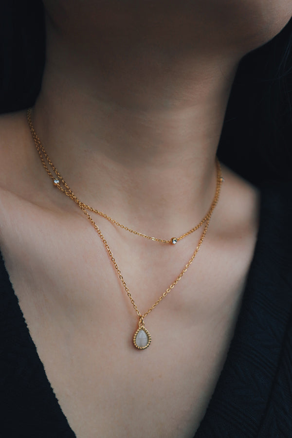 Layered gold necklace set featuring a teardrop-shaped pendant with a mother-of-pearl inlay, paired with a delicate chain adorned with small cubic zirconia stones, worn with a black V-neck top.