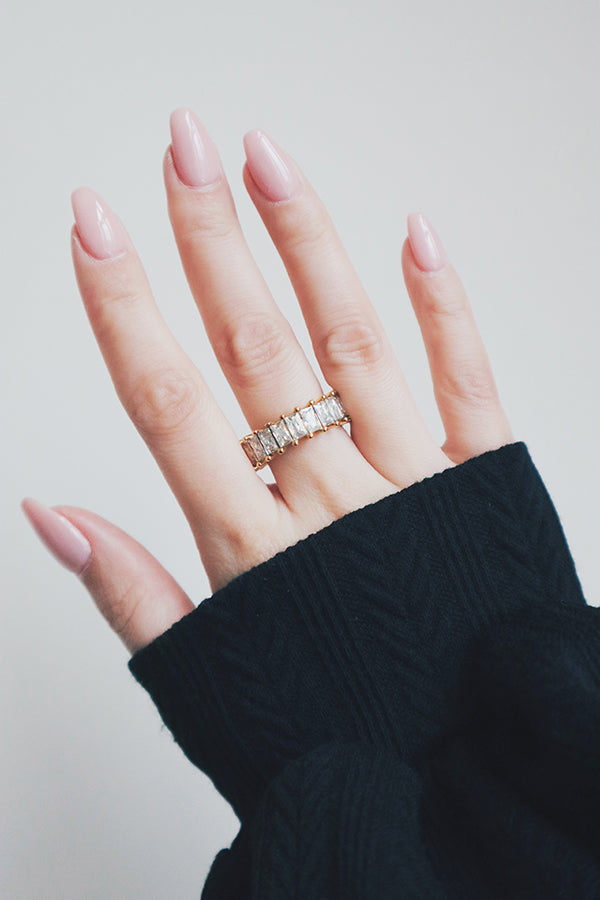 A hand with neatly manicured nails is adorned with a bold gold eternity ring featuring rectangular-cut cubic zirconia stones. The rings large, sparkling stones are set in a sturdy prong setting, creating a luxurious and eye-catching design. The black knit sleeve contrasts with the ring, making it stand out as a statement piece that exudes elegance and sophistication.