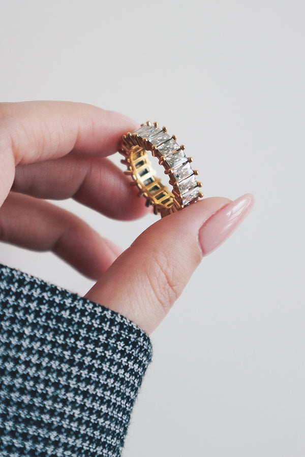 A hand holding a chunky gold eternity ring with rectangular-cut cubic zirconia stones. The ring's intricate design features a series of large, sparkling stones set in a prong setting, creating a timeless and luxurious look. The hand's delicate fingers and neutral manicure further highlight the ring's striking and sophisticated style.