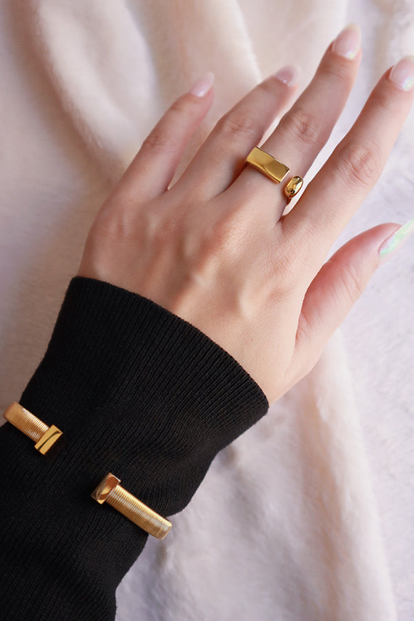 一隻女生的手佩戴一枚潮流金色開口幾何戒指,設計帶有長方形金條和球體,並搭配同款手鍊。