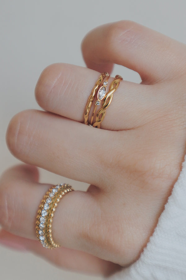 A hand wearing a stack of three dainty gold rings, including twisted bands and a ring with small, clear cubic zirconia stones.