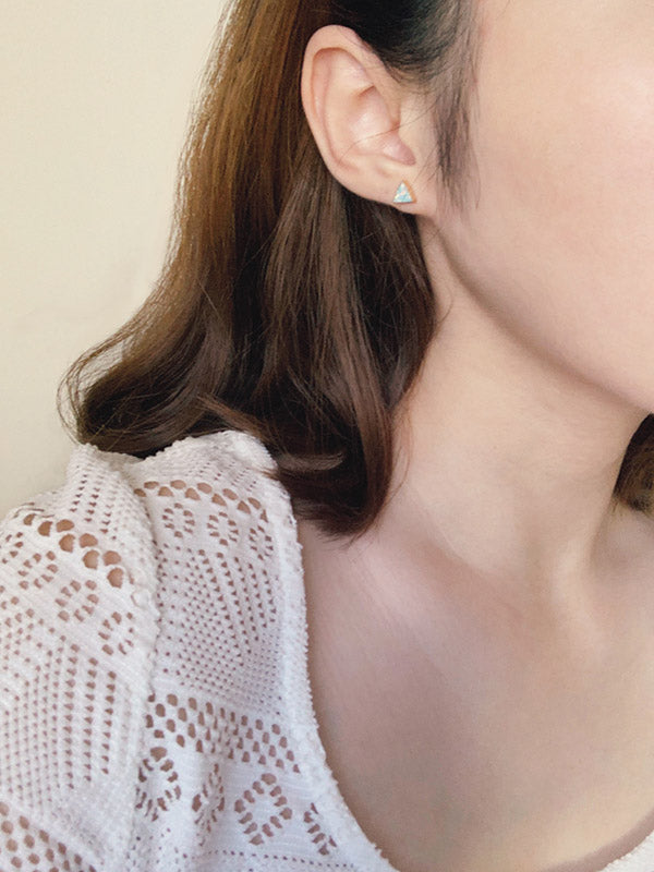 A close-up side profile of a woman's ear, neck, and shoulder, showcasing a small gold-toned triangle stud earring with an iridescent blue opal inlay. The woman has long brown hair and is wearing a white knitted or lace top.