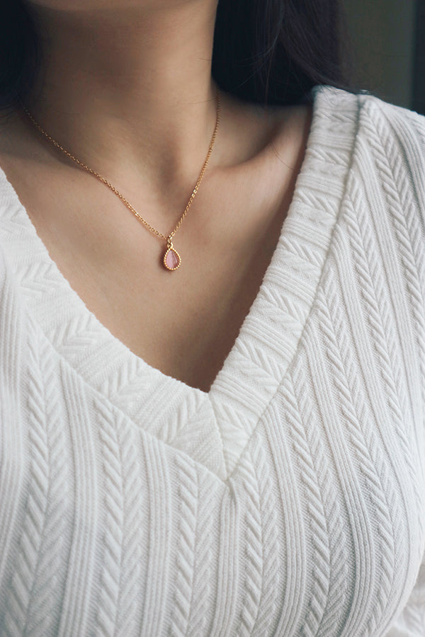 A delicate waterproof necklace featuring a pink cat's eye gemstone in a teardrop-shaped pendant, styled with a white textured V-neck top, perfect for summer.