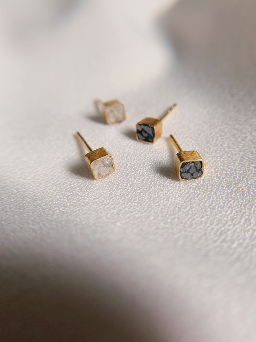 An overhead shot of four gold-toned square stud earrings with crushed stone inlays, two white and two black. They are arranged on a white textured surface, with soft light and shadows.