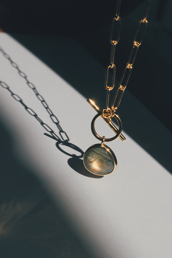 一條女性佩戴的抗過敏金色迴紋針鍊條項鍊,鏈身設計現代簡約,中央配有一個方便開合的T字扣環,下方懸掛著一顆具有迷人虹彩光澤的拉長石墜飾,在陽光下投射出優雅的陰影。