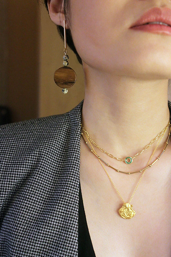 一位女性耳朵和胸部的側面特寫,展示著一枚帶有深色木盤和斑點珠子的長垂墜耳環。這位女性還佩戴著三條疊戴的金色項鍊:一條帶孔雀石環的迴紋針鏈、一條珠狀條紋鏈,以及一條帶有抽象金色吊墜的精緻鏈。這位女性穿著灰黑色格紋西裝外套。