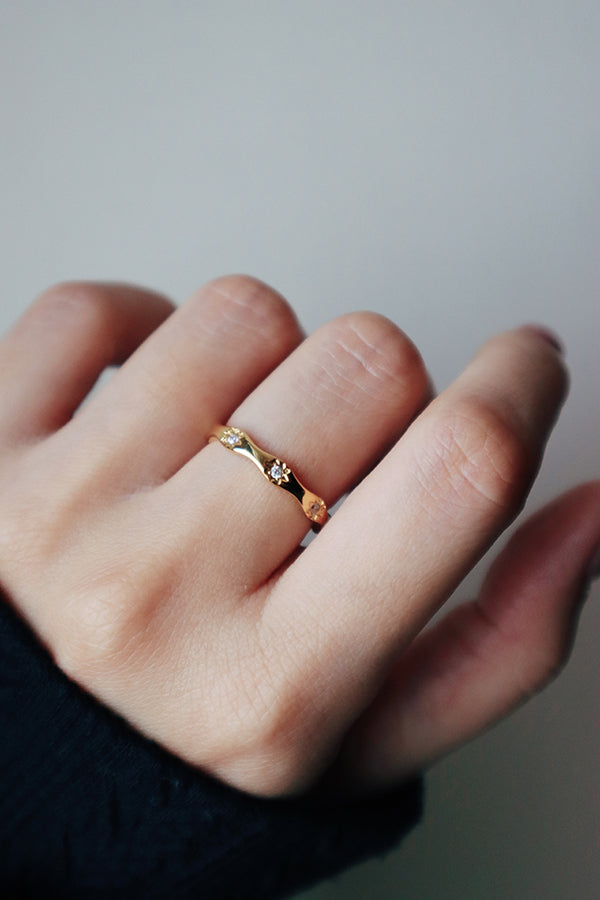 A very close-up shot of a woman's hand, highlighting the Amara Sparkle Ring on her finger. The delicate gold band has a subtle wavy profile with two tiny, embedded white stones. The woman is wearing a black cuff.